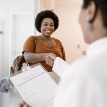 A middle-aged woman enters the doctor's exam room and shakes the hand of a female doctor holding a clipboard of forms.