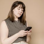 An overweight white adult woman with brunette hair looks down at the smartphone that she is holding with both hands