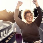 An overweight young woman is cheering with arms overhead after the completion of a physical activity. She is standing on a road, with another female runner behind her.