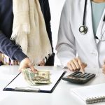 The mid sections of both a woman and a female doctor behind a counter fill the screen. The female patient is putting down cash and the doctor is punching numbers into a calculator.